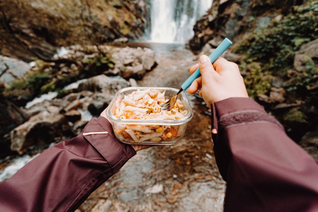 Iemand eet een gevriesdroogde maaltijd uit de verpakking, met een waterval op de achtergrond